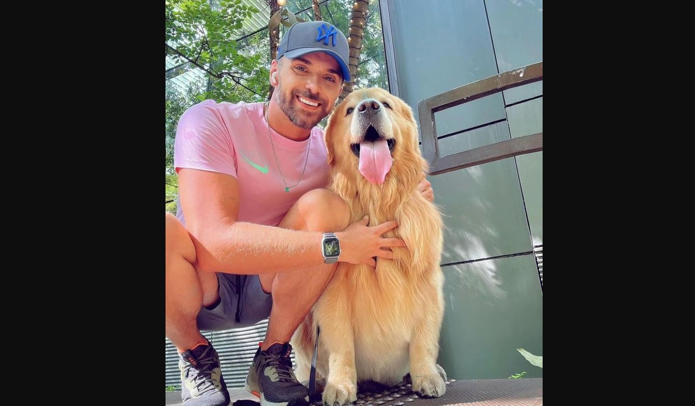 Homem branco com bermuda, camiseta rosa e boné azul está agachado ao lado de cão da raça Golden Retriever